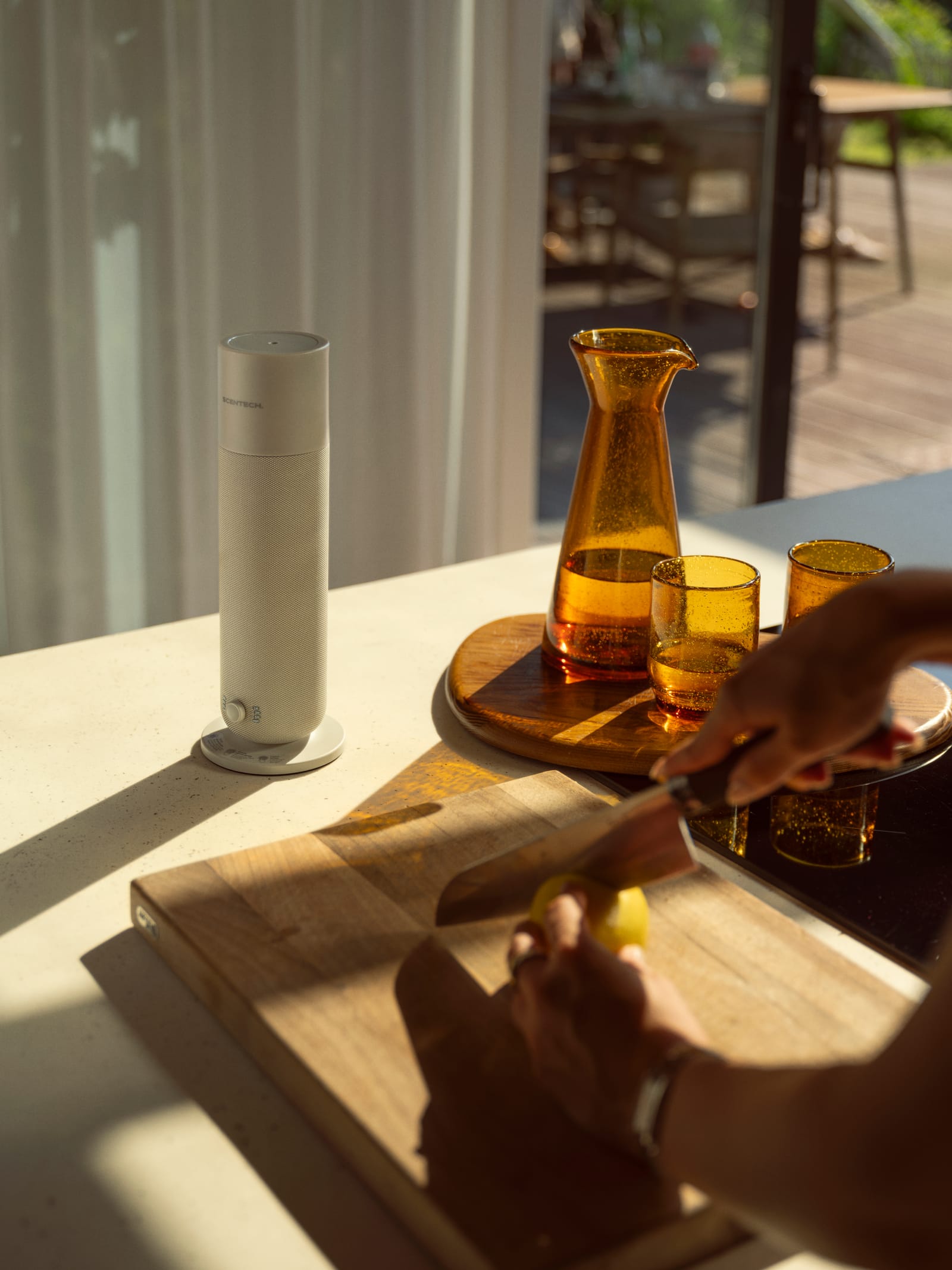 Scentech Smart Aroma Diffuser 2 on a worktop beside an amber carafe while someone slices a lemon